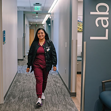 A smiling woman in scrubs walks down a hallway, past a medical lab.