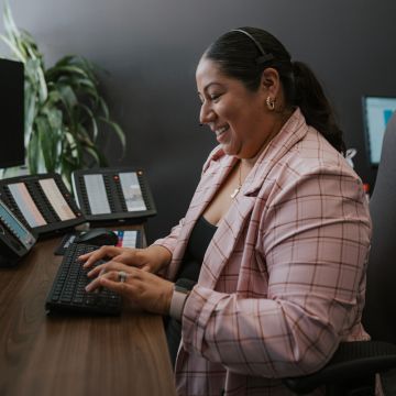 A woman smiling while working at a desk with multiple phone consoles and a keyboard, wearing a headset and a pink checkered blazer.