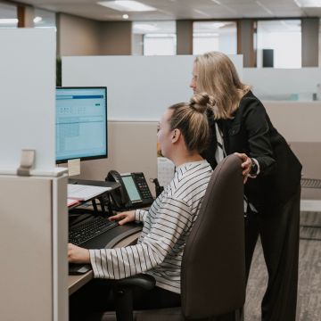 Two women working together in an office cubicle. One person is seated and using a computer, while the other stands beside her, leaning in to provide assistance.